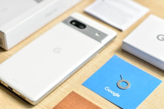 A smartphone with a white back and a metallic strip housing a camera lies on a wooden surface. Nearby, a blue card with a SIM card ejector tool and the word Google is visible, along with various boxes and papers related to the phone.