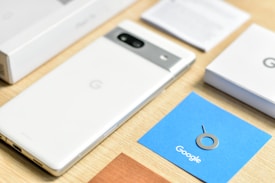 A smartphone with a white back and a metallic strip housing a camera lies on a wooden surface. Nearby, a blue card with a SIM card ejector tool and the word Google is visible, along with various boxes and papers related to the phone.