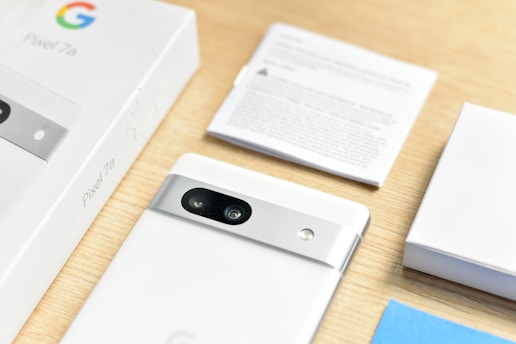 A neatly arranged setup includes a white smartphone partially removed from its box, featuring a dual-camera system and metallic accents on the back. The product name 'Pixel 7a' is visible on the box. Additional packaging materials and a user manual are placed nearby on a light wooden surface.