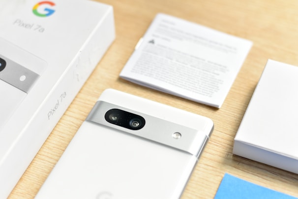 A neatly arranged setup includes a white smartphone partially removed from its box, featuring a dual-camera system and metallic accents on the back. The product name 'Pixel 7a' is visible on the box. Additional packaging materials and a user manual are placed nearby on a light wooden surface.