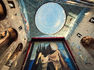 A large, surreal painting depicting a partially abstract human figure with a barren tree on its body is displayed prominently on a tall wall. The scene is framed by an ornate, spherical, glass ceiling above. The surrounding walls feature arches and are decorated with various sculptures and murals.