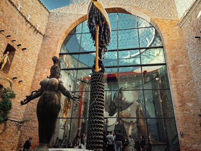 A large, outdoor courtyard area is shown with stone walls and a large glass window reflecting an intricate sculpture. The sculpture features a prominent feminine figure with outstretched arms. There are multiple people present admiring the artwork and the architecture which includes arched windows and ivy-covered walls.