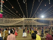 A nighttime gathering with numerous people dressed in traditional attire, dancing or socializing under a canopy of hanging lights. The event appears festive with the lights radiating a warm glow. Decorations can also be seen, including hanging colorful objects on the left side.