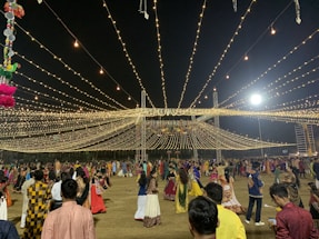 A nighttime gathering with numerous people dressed in traditional attire, dancing or socializing under a canopy of hanging lights. The event appears festive with the lights radiating a warm glow. Decorations can also be seen, including hanging colorful objects on the left side.
