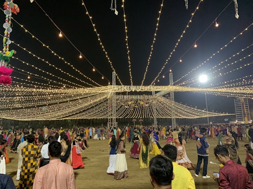 A nighttime gathering with numerous people dressed in traditional attire, dancing or socializing under a canopy of hanging lights. The event appears festive with the lights radiating a warm glow. Decorations can also be seen, including hanging colorful objects on the left side.