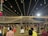 A nighttime gathering with numerous people dressed in traditional attire, dancing or socializing under a canopy of hanging lights. The event appears festive with the lights radiating a warm glow. Decorations can also be seen, including hanging colorful objects on the left side.