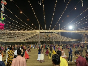 A nighttime gathering with numerous people dressed in traditional attire, dancing or socializing under a canopy of hanging lights. The event appears festive with the lights radiating a warm glow. Decorations can also be seen, including hanging colorful objects on the left side.