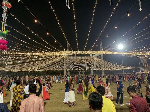 A nighttime gathering with numerous people dressed in traditional attire, dancing or socializing under a canopy of hanging lights. The event appears festive with the lights radiating a warm glow. Decorations can also be seen, including hanging colorful objects on the left side.