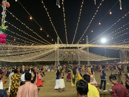 A nighttime gathering with numerous people dressed in traditional attire, dancing or socializing under a canopy of hanging lights. The event appears festive with the lights radiating a warm glow. Decorations can also be seen, including hanging colorful objects on the left side.