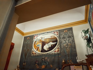 A large oval painting displayed on a wall, featuring surrealistic elements and abstract forms. The artwork is framed with an ornate border and set against a backdrop with intricate designs and shapes. There are decorative objects on a mantelpiece below, including statuettes and figurines. The room's architecture includes a prominent crown molding in a contrasting color.
