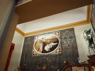A large oval painting displayed on a wall, featuring surrealistic elements and abstract forms. The artwork is framed with an ornate border and set against a backdrop with intricate designs and shapes. There are decorative objects on a mantelpiece below, including statuettes and figurines. The room's architecture includes a prominent crown molding in a contrasting color.
