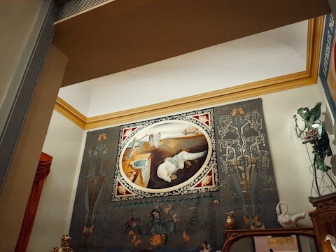 A large oval painting displayed on a wall, featuring surrealistic elements and abstract forms. The artwork is framed with an ornate border and set against a backdrop with intricate designs and shapes. There are decorative objects on a mantelpiece below, including statuettes and figurines. The room's architecture includes a prominent crown molding in a contrasting color.