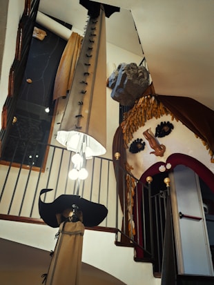 An interior space with an eclectic and surreal design, featuring elements such as oversized lips made of upholstery, a large hat-like sculpture, and a curtain resembling hair or grain. The space is accented by wrought iron railings, a stone-like structure, and positioned lighting.
