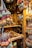 A market stall displaying an array of vibrant and colorful dried fruit and spice garlands. These garlands, featuring oranges, red peppers, green star-shaped accents, and acorns, are neatly arranged and hanging from wooden beams. The stall is rustic, with a wooden backdrop and various small wicker baskets filled with other goods, such as essential oil bottles.