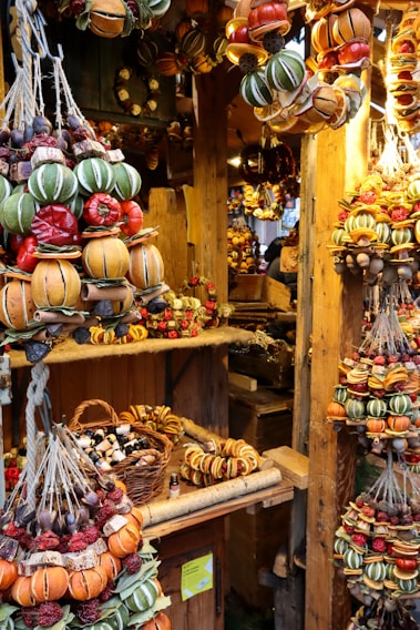 A market stall displaying an array of vibrant and colorful dried fruit and spice garlands. These garlands, featuring oranges, red peppers, green star-shaped accents, and acorns, are neatly arranged and hanging from wooden beams. The stall is rustic, with a wooden backdrop and various small wicker baskets filled with other goods, such as essential oil bottles.