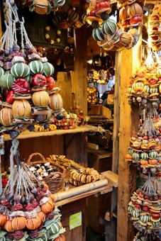 A market stall displaying an array of vibrant and colorful dried fruit and spice garlands. These garlands, featuring oranges, red peppers, green star-shaped accents, and acorns, are neatly arranged and hanging from wooden beams. The stall is rustic, with a wooden backdrop and various small wicker baskets filled with other goods, such as essential oil bottles.