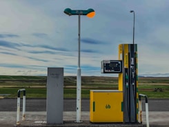 An isolated gas station with a yellow fuel pump labeled for gasoline and diesel stands in a remote landscape. The area around it is open, with a vast expanse of fields in the background and a cloudy sky overhead. The environment appears calm and deserted.