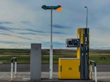 An isolated gas station with a yellow fuel pump labeled for gasoline and diesel stands in a remote landscape. The area around it is open, with a vast expanse of fields in the background and a cloudy sky overhead. The environment appears calm and deserted.