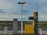 An isolated gas station with a yellow fuel pump labeled for gasoline and diesel stands in a remote landscape. The area around it is open, with a vast expanse of fields in the background and a cloudy sky overhead. The environment appears calm and deserted.