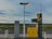 An isolated gas station with a yellow fuel pump labeled for gasoline and diesel stands in a remote landscape. The area around it is open, with a vast expanse of fields in the background and a cloudy sky overhead. The environment appears calm and deserted.