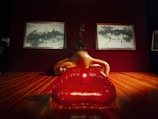 A surreal art installation featuring a large red sofa resembling lips, a structure resembling a human nose above it, and two framed artworks in the background. The room has wooden flooring and a dark, atmospheric lighting.