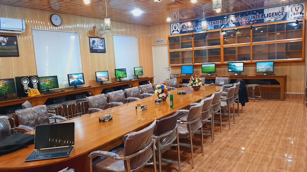 A conference room with a long wooden table surrounded by chairs covered in protective plastic. Multiple computer monitors are placed on tables against the walls, each displaying nature-themed images. The walls are lined with wood paneling, and a sign indicating a center for artificial intelligence is displayed at the front. There are decorative flower arrangements on the table, and the overall setup suggests a professional environment.