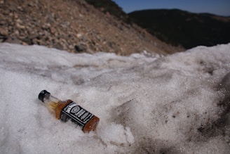 A small bottle of liquor, labeled with a black and white design, lies on a patch of snow on a rocky terrain. The background shows a slope covered in small stones and extends to distant hills under a clear sky.