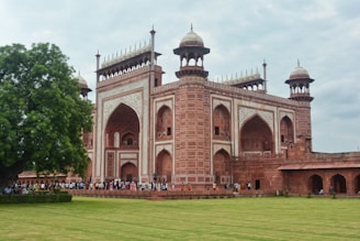 A large, ornate red sandstone gateway with intricate white marble inlay adorns the building. Several symmetrical arches and domed minarets rise from its structure. A lush green lawn and a large tree are present in the foreground, while a line of people is visible entering and leaving the building.