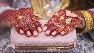 Hands adorned with intricate henna designs and several ornate rings are placed delicately on a shimmering, textured clutch. The background features soft, blurred elements, enhancing the focus on the hands and accessories.