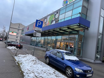A commercial building with large glass panels and signage on the facade, indicating it might be a shopping center or store. There's a parking lot in front with several cars, and a parking sign showing a one-hour limit. The ground is covered with patches of snow, suggesting a cold or winter season. The sky is overcast.