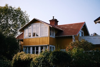 A charming two-story wooden house with a yellow facade and a brown tiled roof. The upper level features large windows, and there is a chimney protruding from the roof. Surrounding the house are lush green trees and bushes, creating a serene and cozy atmosphere.