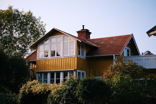 A charming two-story wooden house with a yellow facade and a brown tiled roof. The upper level features large windows, and there is a chimney protruding from the roof. Surrounding the house are lush green trees and bushes, creating a serene and cozy atmosphere.