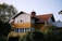 A charming two-story wooden house with a yellow facade and a brown tiled roof. The upper level features large windows, and there is a chimney protruding from the roof. Surrounding the house are lush green trees and bushes, creating a serene and cozy atmosphere.