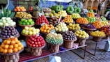A vibrant and colorful display of various fruits arranged in wicker baskets. The assortment includes bananas, pineapples, oranges, grapes, apples, and other tropical and local fruits. The market setting adds a local and fresh vibe, with the fruits stacked neatly to create appealing patterns and textures.