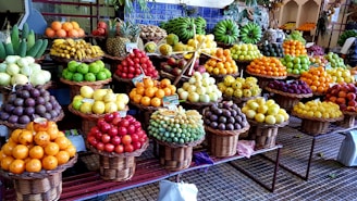 A vibrant and colorful display of various fruits arranged in wicker baskets. The assortment includes bananas, pineapples, oranges, grapes, apples, and other tropical and local fruits. The market setting adds a local and fresh vibe, with the fruits stacked neatly to create appealing patterns and textures.