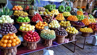 A vibrant and colorful display of various fruits arranged in wicker baskets. The assortment includes bananas, pineapples, oranges, grapes, apples, and other tropical and local fruits. The market setting adds a local and fresh vibe, with the fruits stacked neatly to create appealing patterns and textures.