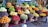 A vibrant and colorful display of various fruits arranged in wicker baskets. The assortment includes bananas, pineapples, oranges, grapes, apples, and other tropical and local fruits. The market setting adds a local and fresh vibe, with the fruits stacked neatly to create appealing patterns and textures.