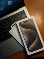 A boxed smartphone is placed on top of a laptop keyboard, with the laptop partially visible. The smartphone box has a sleek design with the image of the phone on the front. The laptop screen shows abstract shapes on a blue background. A wooden surface is noticeable in the background.