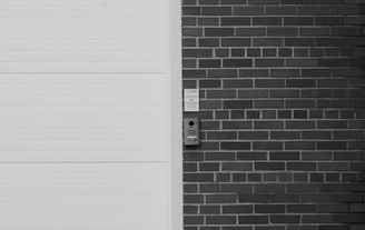 A monochrome image featuring a section of a brick wall with a mounted intercom-like device next to a white, horizontal-lined door. There are visible words above the device indicating 'DELIVERIES AND CUSTOMER PICK-UP'.