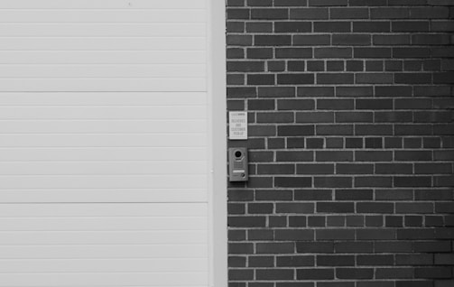 A monochrome image featuring a section of a brick wall with a mounted intercom-like device next to a white, horizontal-lined door. There are visible words above the device indicating 'DELIVERIES AND CUSTOMER PICK-UP'.