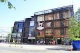 A modern multi-story building with a glass and metal facade, featuring a Dalmatian motif logo and signage for 'FAM TIME' and various stores. People are seen walking near the entrance. The surrounding area includes trees, a large planter, and a traffic cone, indicating a lively urban environment.