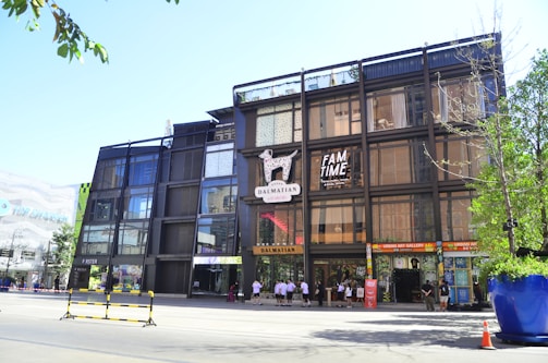A modern multi-story building with a glass and metal facade, featuring a Dalmatian motif logo and signage for 'FAM TIME' and various stores. People are seen walking near the entrance. The surrounding area includes trees, a large planter, and a traffic cone, indicating a lively urban environment.