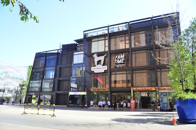 A modern multi-story building with a glass and metal facade, featuring a Dalmatian motif logo and signage for 'FAM TIME' and various stores. People are seen walking near the entrance. The surrounding area includes trees, a large planter, and a traffic cone, indicating a lively urban environment.