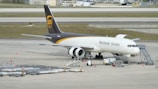A UPS cargo airplane is parked on an airport tarmac with ground support equipment nearby. The plane is white with brown and gold detailing, featuring the logo and the words 'Worldwide Services' on its side. Several orange cones and cargo loading equipment are visible around the plane.