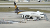 A UPS cargo airplane is parked on an airport tarmac with ground support equipment nearby. The plane is white with brown and gold detailing, featuring the logo and the words 'Worldwide Services' on its side. Several orange cones and cargo loading equipment are visible around the plane.