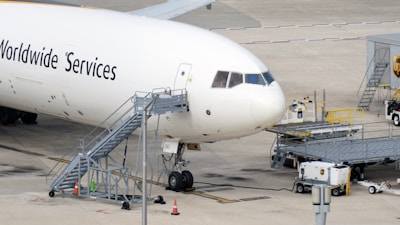 Large commercial aircraft parked on an airport tarmac with a mobile staircase attached to the side. Ground support equipment and vehicles are nearby, along with cargo handling facilities. The words 'Worldwide Services' are visible on the aircraft's fuselage.