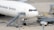 Large commercial aircraft parked on an airport tarmac with a mobile staircase attached to the side. Ground support equipment and vehicles are nearby, along with cargo handling facilities. The words 'Worldwide Services' are visible on the aircraft's fuselage.