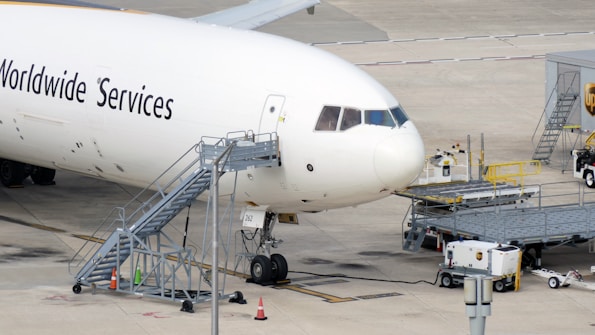 Large commercial aircraft parked on an airport tarmac with a mobile staircase attached to the side. Ground support equipment and vehicles are nearby, along with cargo handling facilities. The words 'Worldwide Services' are visible on the aircraft's fuselage.