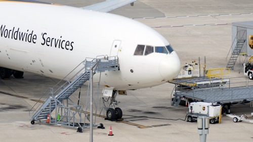 Large commercial aircraft parked on an airport tarmac with a mobile staircase attached to the side. Ground support equipment and vehicles are nearby, along with cargo handling facilities. The words 'Worldwide Services' are visible on the aircraft's fuselage.