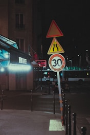 A dimly lit urban street at night features a set of traffic signs, including a speed limit sign showing 30 and a caution sign for narrowing road. A bus appears to be parked in the background, with some neon lights visible on a nearby building. The scene conveys a quiet, possibly deserted city corner.